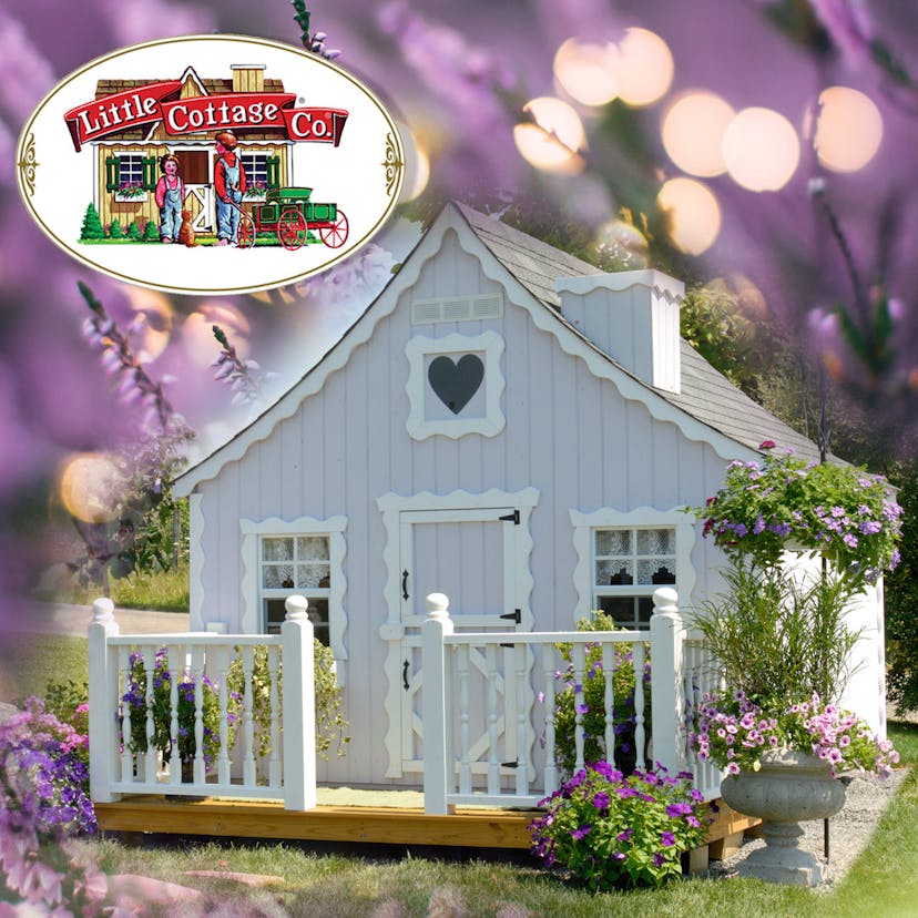 Playhouse with white gingerbread trim, deck and rail, and heart window above small door. Background is purple flowers and lights.